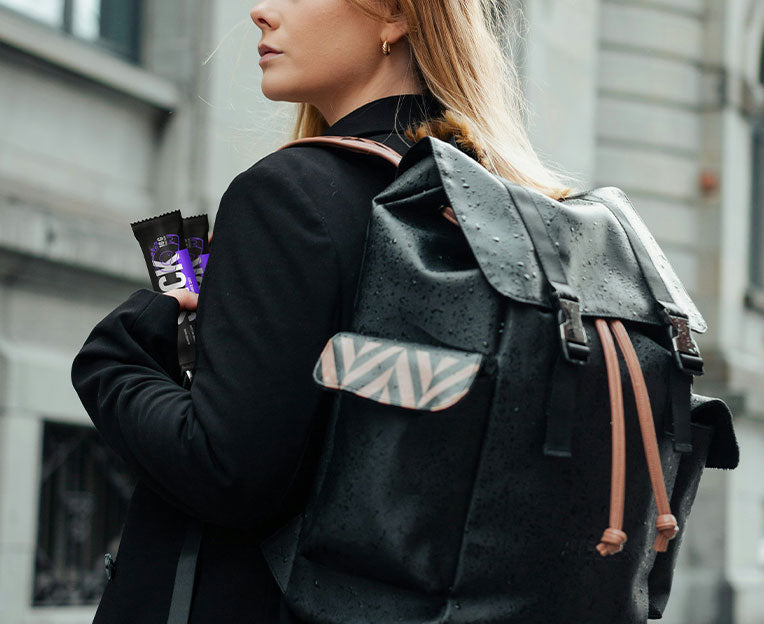 Person holding two SNCK bars wearing a black backpack with a geometric pattern on a blurred city street background