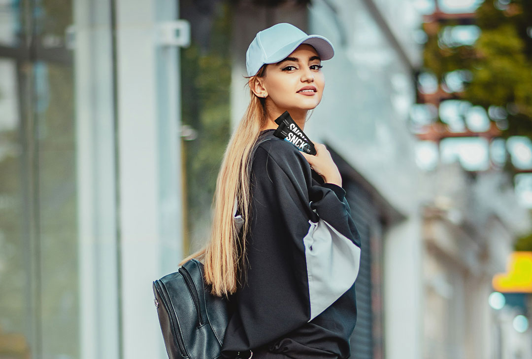 Woman holding two SNCK bars, wearing a light blue cap and black jacket with a blurred city street background